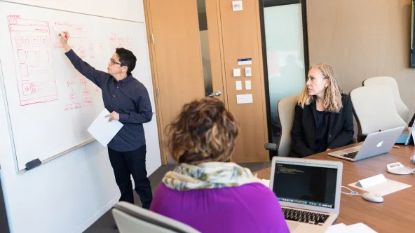 A person writes on a dry erase board while other professionals watch at a board table